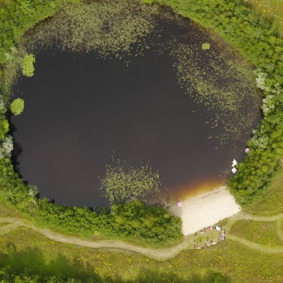 Zwemvijver gezien vanaf boven. Aan de vijver ligt een privéstrand, en het is omringt door bomen en gras waar kronkelende paadjes doorheen lopen.